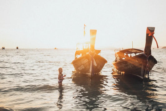 A little girl standing in the sea next to two long-tail boats at sunset in Thailand